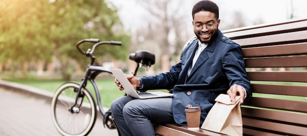 Black man sits on a park bench using his laptop his bicycle nearby the setting is calm and