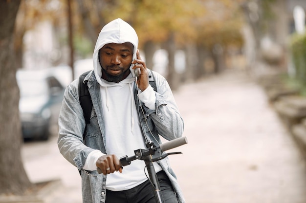 Black man standing on a street with electric scooter. Man wearing white hoodie and black jeans