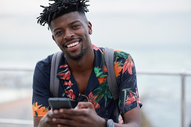 Black man travel and portrait with smartphone and outdoor with communication adventure at beach and backpacking Holiday happy and chat check social media or post with 5g technology and vacation