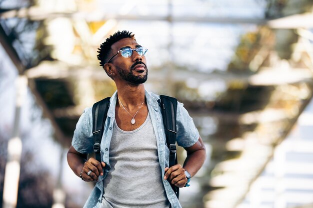 Black man with glasses and backpack walking down the street. Taking the handles of the backpack with his hands.