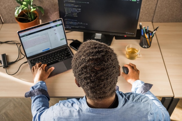 Black Man Writing Lines of Code On Desktop PC With Two Monitors and a Laptop Aside in Stylish Office