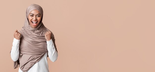 Black muslim woman in headscarf rejoicing success with raised fists