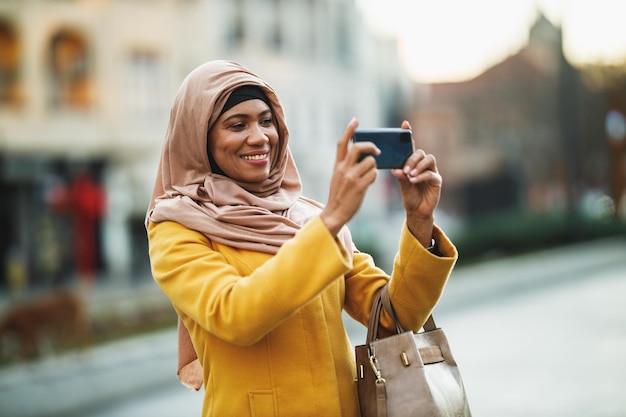 Black Muslim woman wearing a hijab and taking photos by her smartphone standing in urban environment.