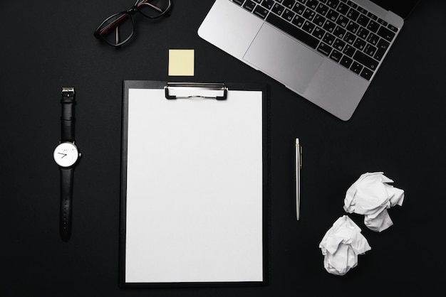 Black office desktop with white sheet of paper with free copy space and pen, laptop, glasses, watch and crumpled paper balls.