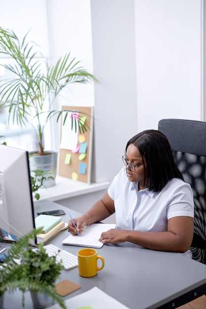 Black office woman sitting at desk working on laptop taking notes at work indoor business schedule c...