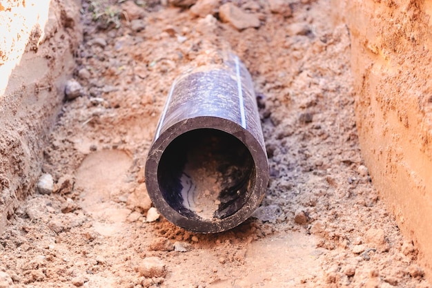 A black plastic pipe with a blue stripe lies in a dug trench to supply water to the house