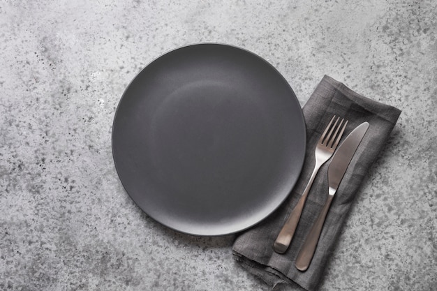 Black plate, fork and knife on grey stone table. Elegance table place setting. Copy space. View from above.