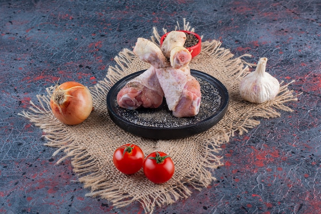 Black plate of raw chicken parts with vegetables on marble surface.