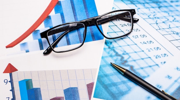 Black-rimmed glasses and a pen in striking charts. Top view of the workplace