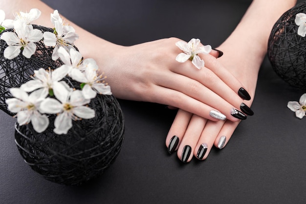 Black and silver manicure with cherry blossom on black background Woman with black nails surrounded with white flowers