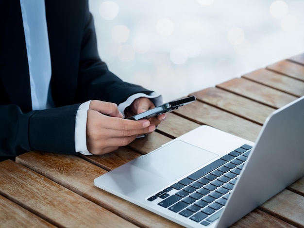 Black smart mobile phone in business woman's hand in suit who holding and using near the notebook computer on desk Texting cybersecurity investment technology and working with two devices concepts