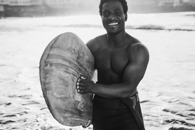 Black surfer man holding vintage surf board on the beach at summer sunset - Focus on face