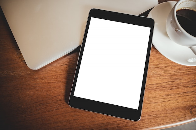 black tablet pc with blank desktop white screen with laptop and coffee cup on wooden table