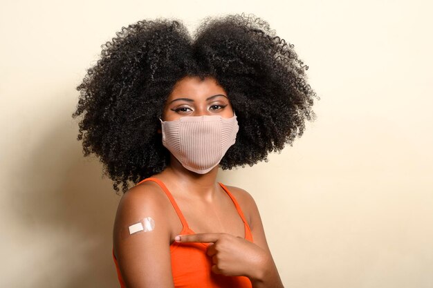 Black teen girl wearing face shield, smiling with her eyes and pointing to the sticker on her arm showing she was vaccinated, covd-19, beige background