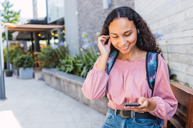 Black university student woman smiling and using her mobile sitting outside the mall in free time