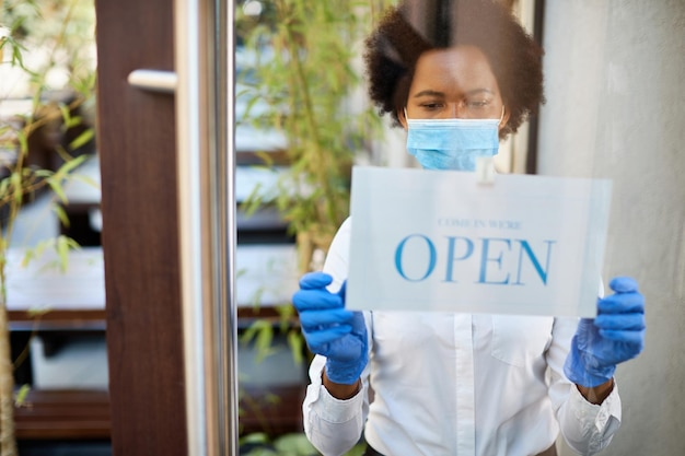 Black waitress with protective face mask and gloves hanging open sign at entrance door