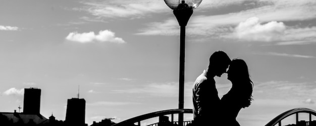 Black and white dark silhouette of couple on the background of big city. Man and woman hugging near the lantern.