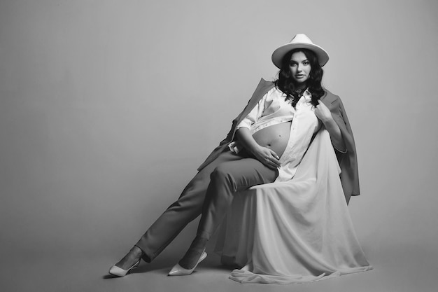 Black and white portrait of brunette pregnant woman in white hat and suit posing on a cyclorama