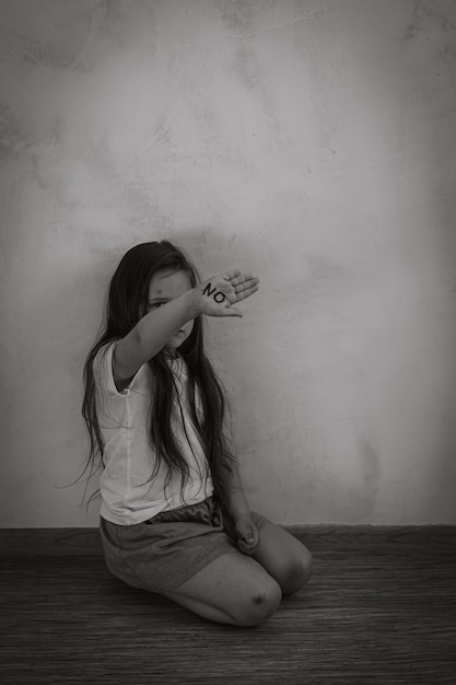 Black and white portrait of little girl sitting on bruised knees showing inscription no on palm Protest against abuse