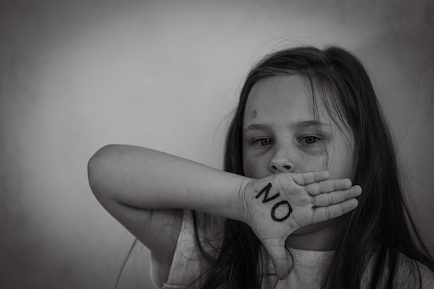 Black and white portrait of little scared bruised girl raising hand to cover mouth showing inscription no on open palm