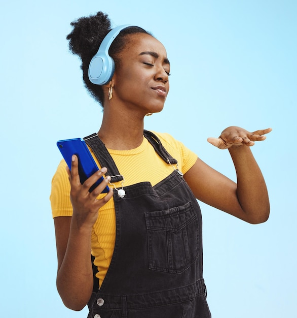 Black woman music and phone in studio with dance dj hand gesture and listening by blue background Gen z girl streaming audio or online radio on internet happy and fashion headphones by backdrop