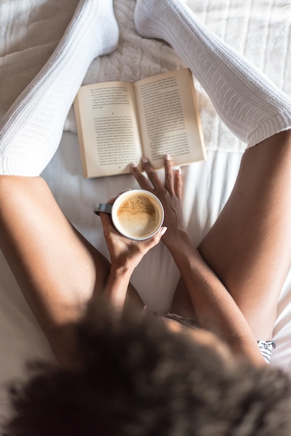 Black woman reading a book and drinking coffee on bed with socks