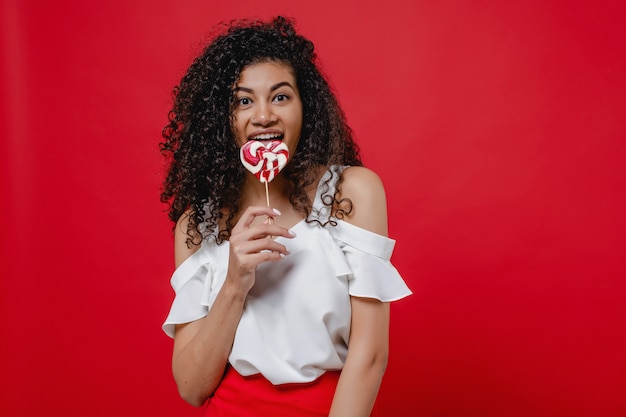 Black woman smiling with heart shaped lollypop candy isolated on red