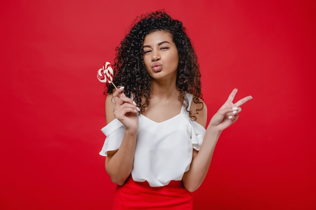 Black woman with heart shaped lollypop candy showing peace gesture on red wall