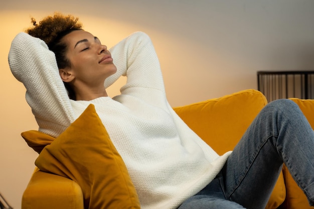 A black young woman is sitting on a comfortable sofa in the living room with her eyes closed and her