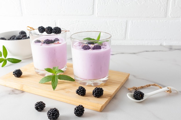 Blackberry smoothie in two glasses on a wooden board with blackberries and mint on a marble white table against the brick wall healthy eating