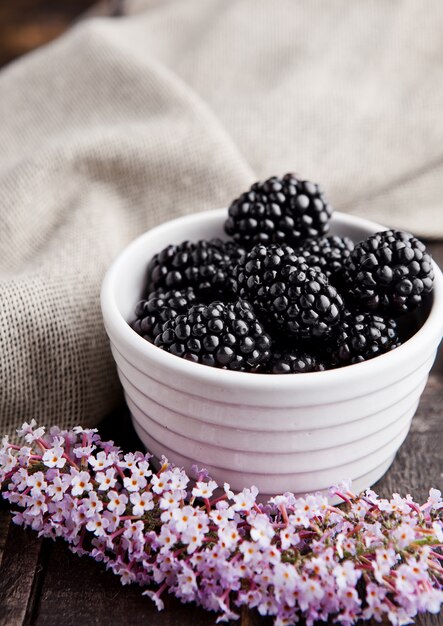 Blackberry in white bowl and flowers on wooden background.