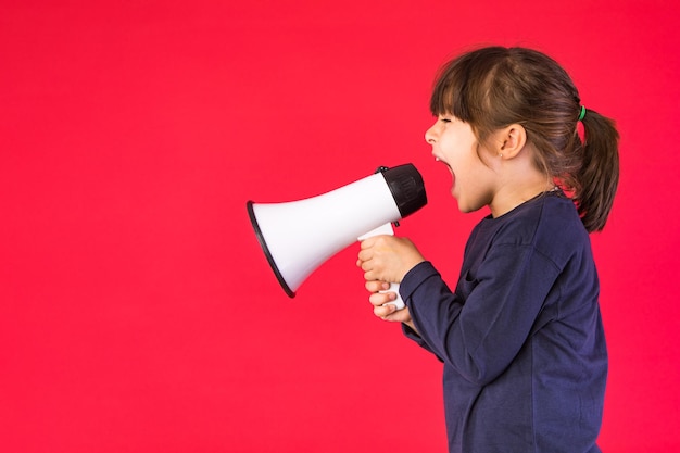 Blackhaired girl in a blue sweater and white pants shouting through a white megaphone on a red background