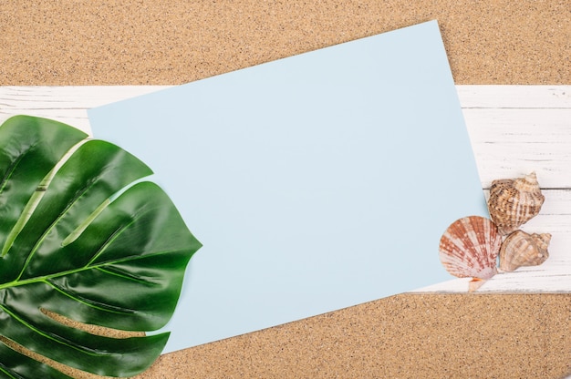 Blank blue sheet of paper, monstera sheet, seashells on white boards on the sand, copy space