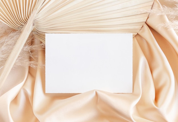 Blank card on beige satin fabric and dried palm leaf close up greeting or wedding mockup