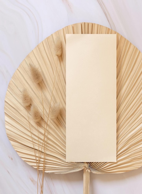 Blank card on dry beige palm leaf near hare's tail grass top view boho wedding mockup