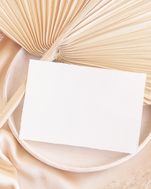 Blank card on plate on dried palm leaf and beige silky fabric close up greeting or wedding mockup