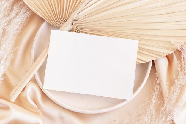 Blank card on plate on dried palm leaf and beige silky fabric close up greeting or wedding mockup