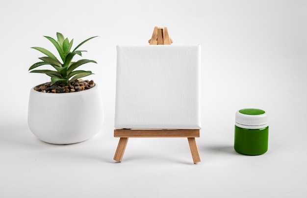 Photo blank clean white canvas on wood mini easel mockup on wood tripod green paint gouache jar and potted plant composition