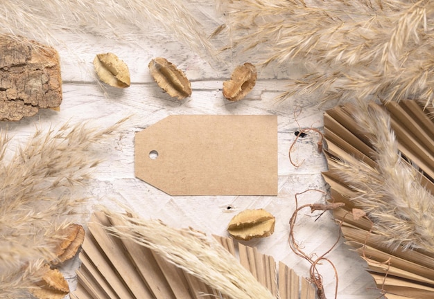 Blank gift tag on white wooden table near dried plants, palm leaves and pampas grass top view. Boho scene with horizontal paper label flat lay, place for text