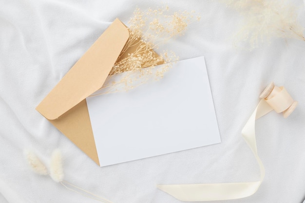 Blank greeting card invitation Mockup 5x7 on envelope with dry flowers and ribbon on white fabric background flat lay mockup