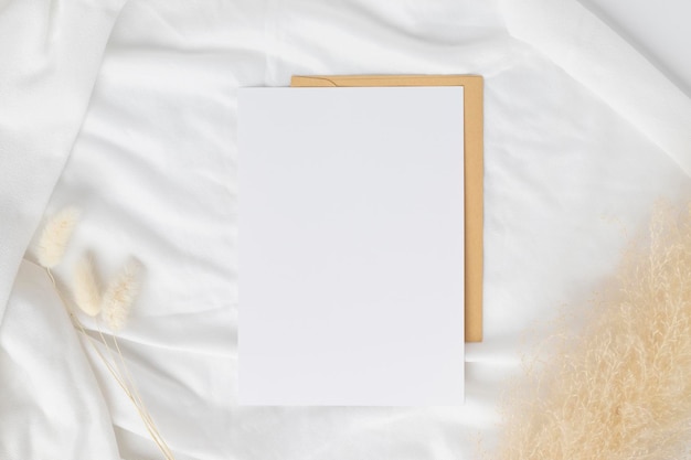 Blank greeting card invitation Mockup on Brown envelope with Dried bunny tails grass on white background Minimal table workplace composition flat lay mockup