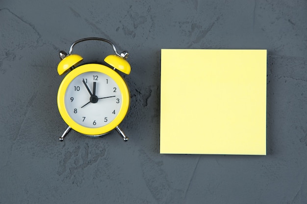 Blank note with alarm clock on gray table, yellow sticky note