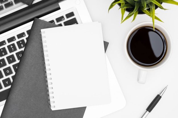 Blank notebook is on top of white office desk table with laptop, coffee cup and office supplies. Top view