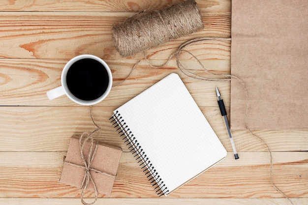 A blank notebook laying on wooden table. It is surrounded by  black pen, cup of coffee, present, rope twine and a paper bag.