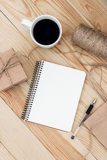 A blank notebook laying on wooden table. It is surrounded by  black pen, cup of coffee, present, rope twine and a paper bag.