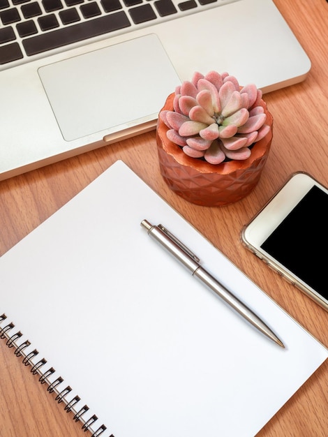 Blank notebook with smartphone laptop computer and succulent plant in copper pot on wooden table background