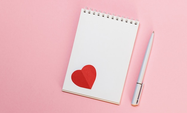 Blank notepad, pen and red paper hearts on pink background. Love message. Valentine's Day and romantic holiday concept. Top view, flat lay with copy space.