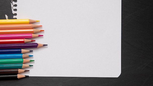 Blank page of a notebook as a mock up next to pencils of different colors on a black chalkboard background