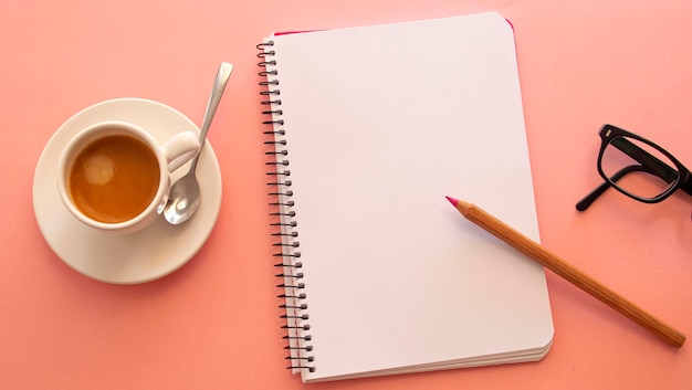 Blank page of a notebook next to a laptop and a white cup with coffee on a light pink background