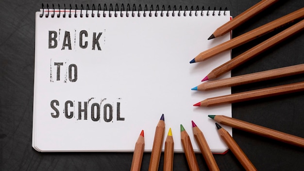 Blank page of a notebook with the phrase quotBack To Schoolquot written in English with worn up letters next to colored pencils on a black chalkboard background